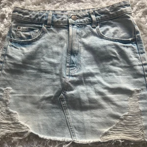 Ljusblå jeanskjol med slitningar - Säljer en ljusblå jeanskjol med råa kanter och slitna detaljer framtill. Kjolen har klassiska jeansfickor både fram och bak samt bälteshällor. Perfekt för dig som gillar en avslappnad och trendig stil. 