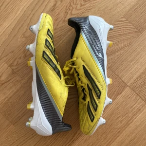 Adidas f50 - Snygga gula adidas fotbollsskor med svarta ränder och vita detaljer. Skorna har snörning och mönstrad ovandel i syntetmaterial. Utrustade med fasta dobbar för bra grepp på planen. Perfekta för dig som vill sticka ut på matchen!
