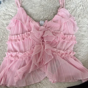 Rosa volangig linnetopp från Fabrika - Supersöt rosa linnetopp från Fabrika med tunna axelband, volanger och rynkade detaljer. Toppens tyg är lätt och plisserat med diskreta ränder och en stor volangrosett framtill. Perfekt för dig som gillar romantisk och lekfull stil.