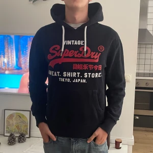 Svart Superdry hoodie med tryck - Svart hoodie från Superdry med stort rött och vitt tryck på bröstet och japanska tecken. Tröjan har huva med vita snören, känguruficka och ribbade muddar. Perfekt för dig som gillar streetwear och vill ha en snygg och bekväm look. OBS! Har en defekt se sista bilden!