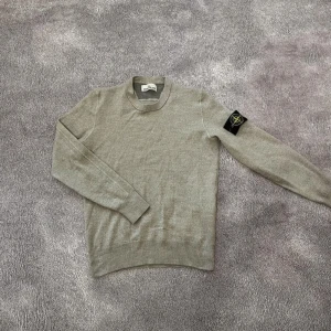 Grå stickad tröja från Stone Island - S med passar bäst i xs. Snygg grå stickad tröja från Stone Island med klassisk rund hals och ikonisk patch på vänster ärm. Tröjan är använd fåtal gånger men skicket är 10/10 skriv för fler bilder