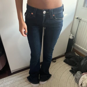Mörkblå True Religion bootcut jeans - Snygga mörkblå jeans från True Religion med bootcut passform. Jeansen har ikoniska bakfickor med lock och silverfärgade knappar, samt kontrastsömmar. Låg midja och klassisk femficksdesign. Perfekta för dig som gillar en tidlös men trendig look. Endast använt vid få tal tillfällen 