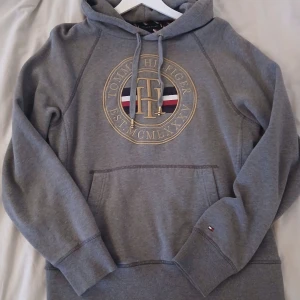 Grå hoodie Tommy Hilfiger S limited edition. - Snygg grå hoodie från Tommy Hilfiger med stort broderat TH-emblem och gulddetaljer på bröstet. Klassisk känguruficka, snörning i huvan och små broderade loggor. Mjuk insida och långärmad modell, perfekt för chill dagar. Nypris 1700kr.