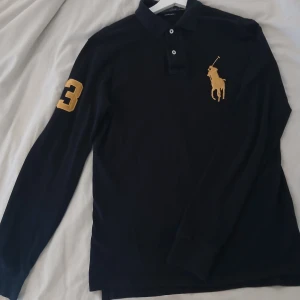 Svart pikétröja Polo Ralph Lauren - Svart långärmad pikétröja från Polo Ralph Lauren i custom slim fit. Stort gult broderat polospelare-logo på bröstet och gul siffra 3 på ärmen. Klassisk krage med knappar och tillverkad i bomull. Perfekt för dig som gillar sportig och stilren look.