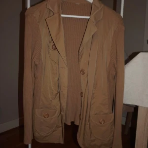 Beige cardigan jacka med knappar - Snygg beige cardigan jacka med ribbade ärmar och två stora fickor framtill. Jackan har knappar och en klassisk krage, perfekt för dig som gillar en chill och stilren look. Materialmix med stickad känsla på ärmar och rygg samt slätare tyg framtill.