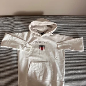 Hoddie GANT Vit - 176 CM/16År - Vit hoodie från GANT med broderad logotyp och sköld på bröstet. Klassisk känguruficka framtill, huva och långa ärmar. Mjuk och skön i bomull, perfekt för en chill och stilren look. Använd någon gång bara. Jätte bra skick. Pris kan diskuteras vid snabbaffär.