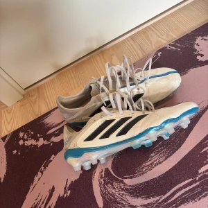 Vita Adidas fotbollsskor med blå detaljer (copa pure III) - Säljer ett par vita Adidas fotbollsskor med svarta ränder och blå detaljer längs sulan. Skorna har snörning och är designade för gräsplan med fasta dobbar. Snygg och sportig look som sticker ut på planen.