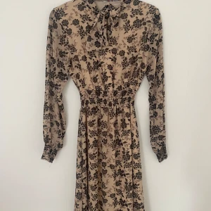 Beige blommig långklänning med rosett - Supersnygg beige långklänning med svart blommigt mönster och markerad midja. Klänningen har långa ärmar med smock vid ärmslut och en söt rosett vid halsen. Perfekt för dig som gillar romantisk och trendig stil.