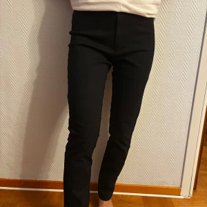 Svarta kostymbyxor från Mango - Svarta kostymbyxor från Mango Basics i klassisk modell med hög midja och smal passform. Byxorna har diskreta bakfickor med knapp och är perfekta för en clean och stilren look. Materialet känns mjukt och stretchigt för extra komfort.