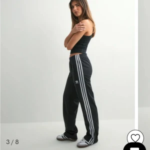 Svarta Adidas byxor med vita ränder - Klassiska svarta Adidas byxor med tre vita ränder längs sidorna och broderad logga på låret. Byxorna har elastisk midja och rak passform, perfekta för en sportig och avslappnad stil.