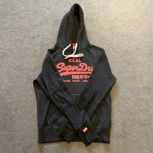 Svart Superdry hoodie med röd logga - Svart hoodie från Superdry med stor röd logga och japansk text på bröstet. Tröjan har huva med vit snörning, känguruficka och ribbade muddar. Perfekt för dig som vill ha en snygg och bekväm look. Tröjan har en liten lagning på baksidan! Men den syns knappt