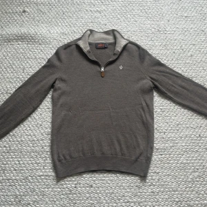 Grå merinoulltröja från Morris - Snygg grå långärmad tröja från Morris i 100% merinoull. Halv dragkedja med läderdetalj och broderad logga på bröstet. Storlek S men passar mer som xs då den har krympt lite. Perfekt för dig som gillar stilrena och klassiska plagg med en modern touch.