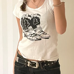 Vit Monki t-shirt med cowboy boots - Vit t-shirt från Monki med svart tryck av cowboy boots på bröstet. Modellen är klassisk med rund hals och korta ärmar. T-shirten är i mjuk bomull och har en normal passform. Perfekt för dig som gillar coola statement-plagg.