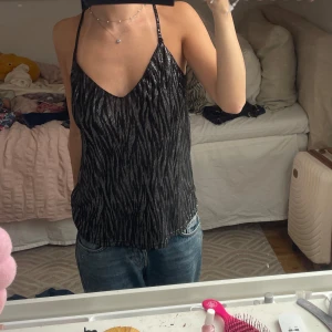 Glittrig svart linne med zebramönster - Säljer ett glittrigt svart linne med tunna axelband och v-ringning både fram och bak. Tyget har ett snyggt zebramönster i silver som ger en cool och trendig look. Perfekt för dig som vill sticka ut lite extra. Passar till jeans eller kjol. 