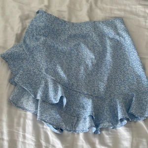 Blå blommig volangkjol - Supersöt blå kortkjol med vitt blommönster och volangkant. Kjolen har en omlott-look och är perfekt för varma dagar. Lätt och luftig känsla, passar dig som gillar en feminin och trendig stil.