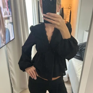 Svart blus med v-ringning och volang - Säljer en svart blus med djup v-ringning och spetsdetaljer längs urringningen. Blusen har långa puffärmar med resår vid handlederna och en lös, flowy passform. Volangdetaljer vid axlarna ger en extra touch. Perfekt för dig som gillar stilrena och trendiga plagg.