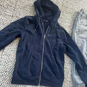 Marinblå hoodie med dragkedja från NEXT - Mysig marinblå hoodie från Next med huva och snörning, hel dragkedja framtill och två stora fickor. Perfekt för chill dagar eller när du vill ha en enkel och stilren look. Tillverkad i mjuk bomullsmix för extra komfort.