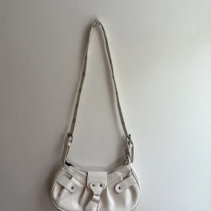 Vit axelväska i croco-imitation - Snygg vit axelväska i croco-imitation med justerbart axelband med metallöglor. Väskan har två fickor framtill med knäppning och en stilren, avlång form. Perfekt accessoar för att lyfta din outfit.