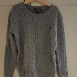 Grå kabelstickad tröja Polo Ralph Lauren - Kabelstickad grå tröja från Polo Ralph Lauren i klassisk modell med rund halsringning och diskret broderad logga på bröstet. Långärmad och ribbade muddar för en snygg passform. Perfekt för lager-på-lager under kyliga dagar.