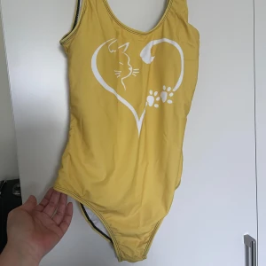 Gul baddräkt med kattmotiv - Supersöt gul baddräkt med vitt tryck av en katt och tassar som bildar ett hjärta på framsidan. Klassisk modell med breda axelband och hel rygg. Perfekt för strand eller poolhäng och har vit insida för extra komfort.Helt ny