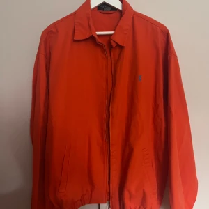 Orange bomberjacka från Yves Saint Laurent - Snygg orange bomberjacka från Yves Saint Laurent med klassisk krage, dragkedja och två fickor framtill. Jackan har blå YSL-brodyr på bröstet och elastisk nederkant. Perfekt för dig som vill sticka ut med en ikonisk och stilren jacka.