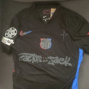 FC Barcelona x Travis scott Pedri tröja - Exklusiv svart FC Barcelona x Travis scott matchtröja med Pedri #8 på ryggen. Tröjan har korta ärmar, broderat klubbmärke med blå och röd kant, Cactus Jack-grafik framtill och officiella UEFA Foundation och Champions League-patchar på ärmarna. Materialet är lätt och andas. Storleken är M men den sitter lite tight