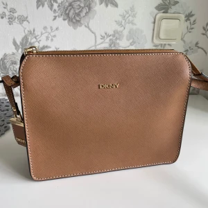 Brun axelväska från DKNY i skinn - Snygg brun axelväska från DKNY i stilren, fyrkantig modell med guldfärgade detaljer och logga framtill. Väskan har justerbar axelrem, dragkedja upptill och ett smidigt ytterfack på baksidan. Tillverkad i strukturerat skinn med kontrastsömmar.