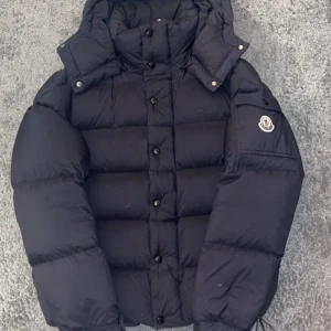 Svart Moncler dunjacka med huva - Säljer en svart Moncler dunjacka med huva och klassisk logga på ärmen. Jackan är quiltad, har knappar framtill och dragsko nertill. Insidan har ett coolt serietryck och mjukt foder. Perfekt för kalla dagar när du vill hålla stilen.