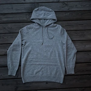 Grå kashmir hoodie med huva - Mysig grå hoodie i kashmir med huva och snörning. Tröjan har långa ärmar och ribbade muddar vid ärmslut och nederkant. Perfekt för dig som gillar en enkel och stilren look.