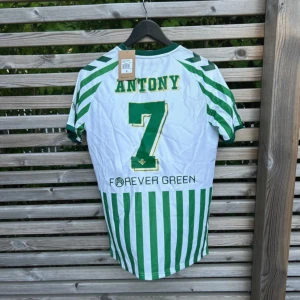 Real Betis Antony #7 matchtröja S - Säljer en officiell Real Betis fotbollströja från Hummel i storlek S. Tröjan är vit med gröna vertikala ränder, gröna detaljer och tryck med Antony #7 på ryggen. Klassisk rund hals och klubbmärke på bröstet. Materialet är lätt och andas, perfekt för match eller träning.