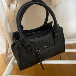 Svart handväska med dragkedja - Snygg svart handväska i skinnimitation med två rejäla handtag och coola sömdetaljer. Väskan har en dragkedja upptill och en mindre ficka med dragkedja framtill, perfekt för småprylar. Stilren och passar till det mesta. Använd en gång!