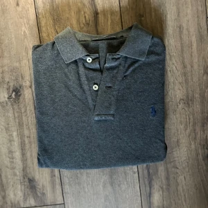 Mörkgrå långärmad piké från Polo Ralph Lauren - Säljer en mörkgrå långärmad pikétröja från Polo Ralph Lauren. Klassisk krage, knäppning med två knappar och broderad blå logga på bröstet. Tröjan är i mjuk bomull och har ribbade muddar vid ärmsluten. Perfekt för en clean och stilren look.