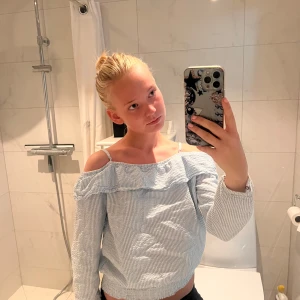 Randig offshoulder blus med volang - Superfin blus med smala blå och vita ränder, offshoulder-modell och volangdetalj längs överdelen. Långärmad med resår i midjan för en snygg passform. Perfekt för vår och sommar när du vill ha en chill men ändå söt look.