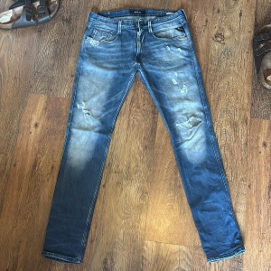 Replay Anbass 33/34 - Säljer ett par Replay Anbass jeans i blå tvätt med slitningar och distressed detaljer. Jeansen har normal passform, raka ben och klassisk femficksdesign. Materialet är mjukt denim i bomull med snyggt slitna partier och Replay-logga på fickan.