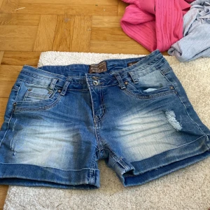 Blå jeansshorts från Monday Denim - Snygga blå jeansshorts från Monday Denim med slitna detaljer och ljusa partier framtill. Klassisk femficksmodell med bälteshällor och uppvikta benslut. Perfekta för sommardagar och enkel att matcha med allt. 