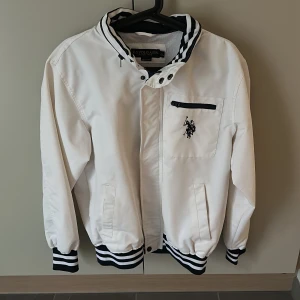 Vit vindjacka från U.S. Polo Assn XL/M - Snygg vit vindjacka från U.S. Polo Assn. i storlek XL men är mer som en medium. Jackan har svarta och vita ränder på krage och muddar, dragkedja och knappstängning, samt en bröstficka med svart dragkedja och broderad logga samt en inbyggd luva. Perfekt för dig som gillar sportig och clean stil.