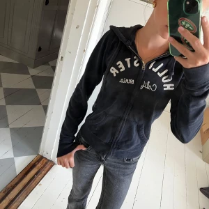 Marinblå zip-hoodie från Hollister - Mysig marinblå hoodie från Hollister med broderad logga och text på bröstet. Jackan har huva med snörning, dragkedja framtill och två fickor. Perfekt för chill dagar eller när du vill ha en avslappnad look.