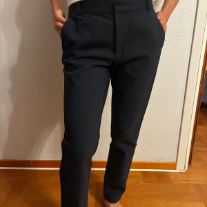 Mörkblå kostymbyxor Stradivarius 34 - Snygga mörkblå kostymbyxor från Stradivarius i storlek 34. Byxorna har raka ben, hög midja och medföljande svart bälte med gulddetalj. Perfekta för en clean och stilren look. Materialet är mjukt och följsamt, med fickor fram och bak.