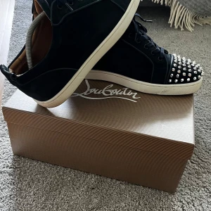 Mörkblå Louboutin sneakers med nitar - Svarta sneakers från Christian Louboutin i mocka med vita sulor och ikoniska röda undersidor. Tåpartiet är täckt av silvriga nitar 