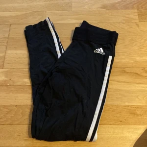 Svarta Adidas mjukisbyxor med ränder - Svarta mjukisbyxor från Adidas med klassiska vita ränder längs sidorna och Adidas-logga på låret. Byxorna har resår i midjan och är perfekta för chill eller träning. Snygg och sportig look som alltid funkar.