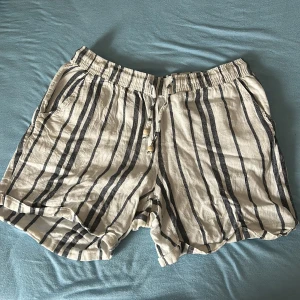 Randiga linneshorts med snörning - Snygga beige shorts i linne med svarta vertikala ränder och avslappnad passform. De har elastisk midja och snörning med träpärlor framtill. Perfekta för varma dagar när du vill ha en chill och stilren look.