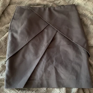 Mörkgrå omlottkjol i kort modell - Snygg mörkgrå kortkjol med omlott-design och stilrena linjer. Kjolen har en modern look och passar perfekt till en trendig outfit. Enkel att matcha med både sneakers och boots.