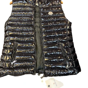 Svart dunväst  - Snygg svart dunväst  med glansig finish och horisontella quiltade sömmar. Västen har två dragkedjefickor framtill, hög krage. Perfekt att slänga över en hoodie för en cool look.
