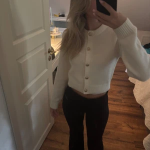 Vit croppad kofta med guldknappar - Superfin vit croppad kofta med snygga guldknappar framtill. Tröjan har rund halsringning, ribbade muddar och är långärmad. Perfekt att styla med höga jeans eller kjol för en clean look.