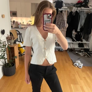 Vit v-ringad blus med guldknappar - Säljer en stilren vit blus med v-ringning och korta ärmar. Blusen har tre snygga guldknappar framtill och en liten slits nertill som ger en modern touch. Passformen är croppad och lätt figursydd, perfekt till svarta byxor eller jeans.