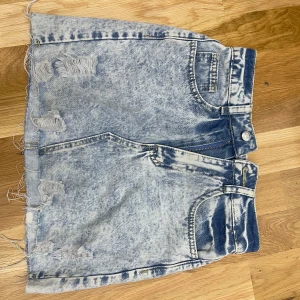 Acid wash jeanskjol med slitningar - Cool ljusblå jeanskjol i acid wash med slitna detaljer och fransig nederkant. Klassisk femficksmodell med dragkedja och knapp framtill. Perfekt för dig som gillar en edgy och avslappnad stil.