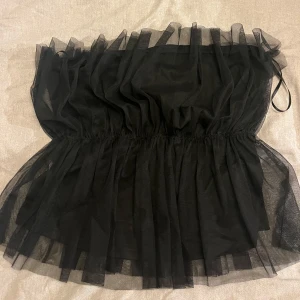 Svart tubtopp i tyll från Vero Moda - Säljer en svart tubtopp från Vero Moda i luftig tyll med volangdetaljer upptill och nertill. Toppen är bandeau-modell och har en snygg, puffig look som ger extra edge till outfiten. Perfekt för dig som vill sticka ut med en cool och trendig stil💕💕