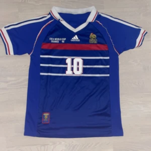 Frankrike Zidane VM 98 Adidas tröja - Klassisk blå fotbollströja från Adidas, Frankrikes landslag VM 1998 med Zidane och nummer 10 på ryggen. Tröjan har vita och röda detaljer, V-ringad krage och korta ärmar. Materialet är lätt och andas, perfekt för fotboll eller samling. Storlek M men hyfsat stor i storlek.
