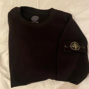 Svart Stone Island sweatshirt - Svart sweatshirt från Stone Island med klassisk rund hals och ikonisk logotyp-patch på vänster ärm. Tröjan är i mjuk bomull och har en clean, stilren look som passar perfekt till streetwear. Ribbstickade muddar vid hals, ärmslut och nederkant.
