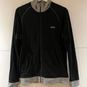 Svart overshirt från BOSS med dragkedja - Svart overshirt från BOSS med grå detaljer vid krage, ärmslut och nederkant. Jackan har hel dragkedja framtill, fickor och diskret BOSS-logga på bröstet. Perfekt för en avslappnad och stilren look.
